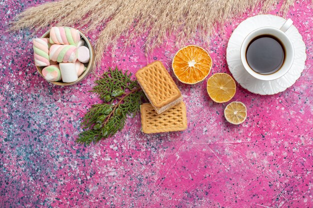 Vista superior de deliciosas galletas waffle con taza de té y malvaviscos en superficie rosa