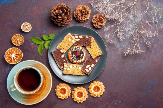 Vista superior de deliciosas rebanadas de pastel con poco de galleta y una taza de té en el fondo oscuro galleta galleta postre pastel té dulce