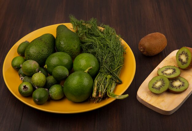 Vista superior de deliciosas rodajas de kiwi en una tabla de cocina de madera con aguacates feijoas y limas en una placa amarilla en una pared de madera