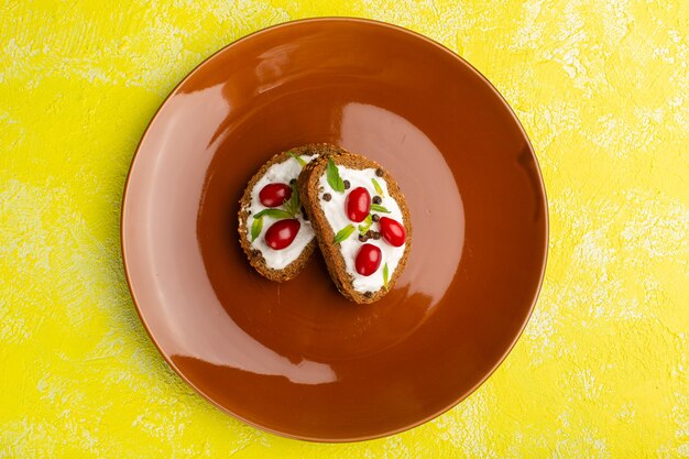 Vista superior de deliciosas tostadas de pan con crema agria y cornejos dentro de la placa marrón en el escritorio amarillo