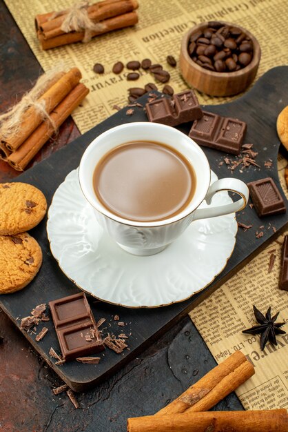 Vista superior de un delicioso café en una taza blanca sobre una tabla de cortar de madera en un periódico antiguo, galletas, canela, limas, barras de chocolate