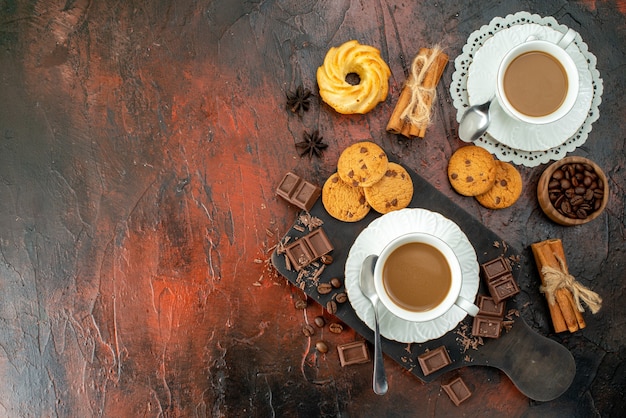 Vista superior del delicioso café en tazas blancas en la tabla de cortar de madera galletas canela limas barras de chocolate en el lado izquierdo sobre fondo de colores mezclados