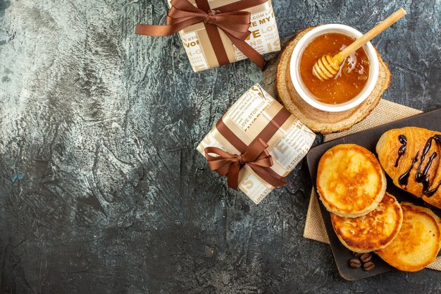 Vista superior del delicioso desayuno con panqueques en la tabla de cortar de madera miel hermosas cajas de regalo en superficie oscura