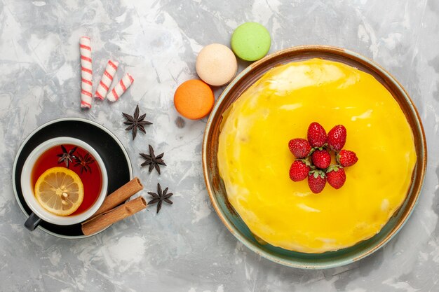 Vista superior delicioso pastel afrutado con macarons de jarabe amarillo y una taza de té en la superficie blanca galleta de pastel dulce azúcar hornear galleta de té