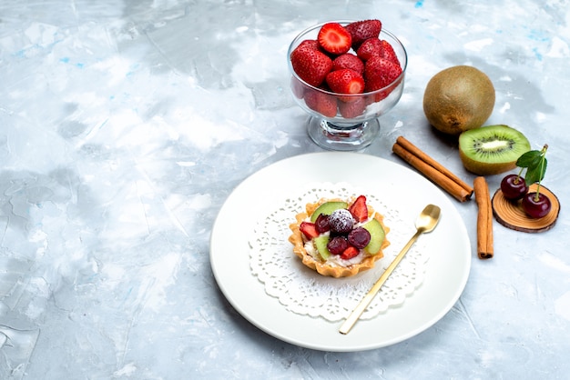 Una vista superior del delicioso pastel con crema y frutas dentro de un plato blanco junto con frutas frescas canela en el escritorio azul grisáceo pastel de frutas azúcar