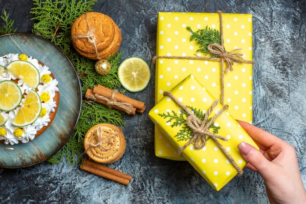 Vista superior del delicioso pastel cremoso y ramas de abeto limón canela limas mano sosteniendo una de las cajas de regalo amarillas sobre fondo oscuro