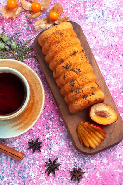 Vista superior delicioso pastel dulce y delicioso con ciruelas canela y taza de té sobre fondo rosa.