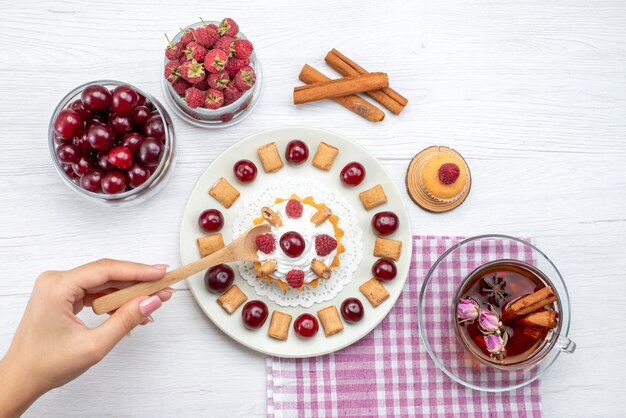 Vista superior del delicioso pastel con frambuesas, cerezas y poco de té de galleta canela en el escritorio de luz, té de crema de frutas y bayas