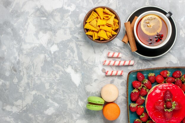 Vista superior delicioso pastel de fresa con fresas rojas frescas taza de té y macarons sobre fondo blanco.