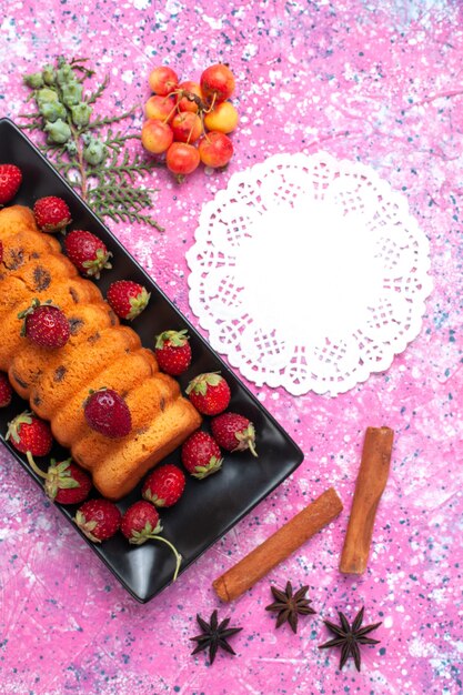 Vista superior delicioso pastel horneado dentro de un molde para pastel negro con fresas rojas frescas y canela en el escritorio rosa.