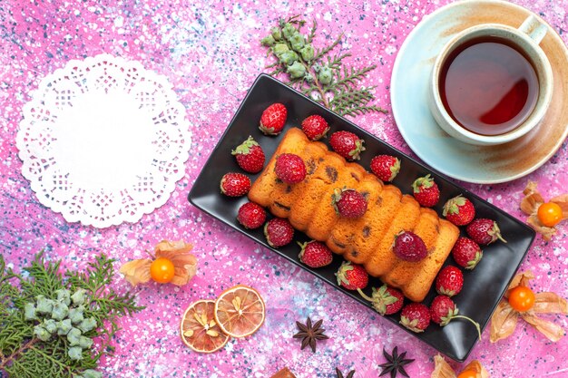 Vista superior delicioso pastel horneado dentro de un molde para pastel negro con fresas rojas frescas y una taza de té en el escritorio rosa.