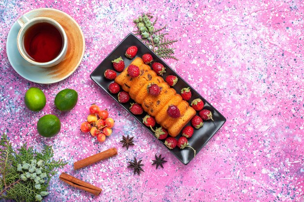 Vista superior delicioso pastel horneado dentro de un molde para pastel negro con fresas rojas frescas té canela y limones en el escritorio rosa.