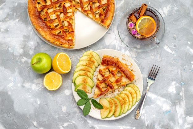 Una vista superior del delicioso pastel de manzana dentro de la placa con té y manzana verde fresca en el escritorio gris pastel de azúcar