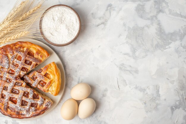 Vista superior del delicioso pastel de mermelada en un plato blanco, huevos con picos de harina en una olla pequeña sobre una mesa blanca con espacio libre