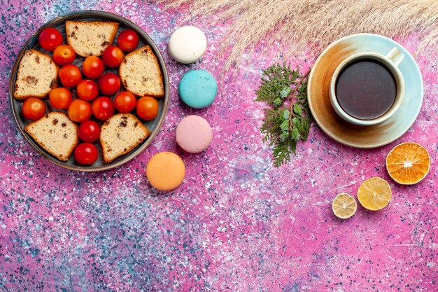 Vista superior del delicioso pastel en rodajas con macarons de ciruelas frescas agrias y una taza de té en el escritorio rosa