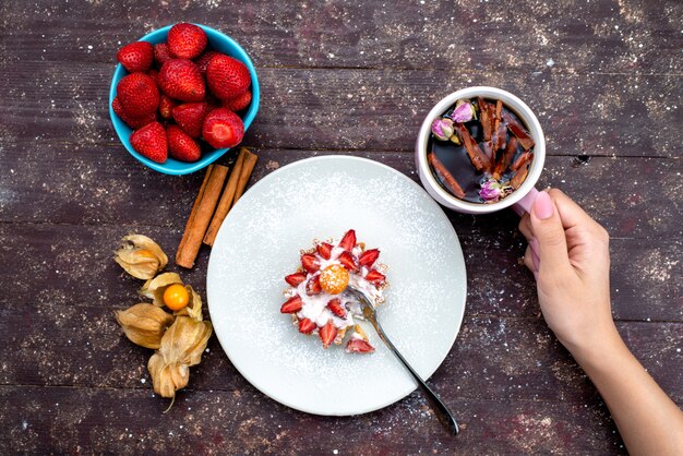 Una vista superior del delicioso pastelito con crema y frutas frescas en rodajas junto con canela y fresas en el escritorio marrón, pastel de frutas, té de galletas