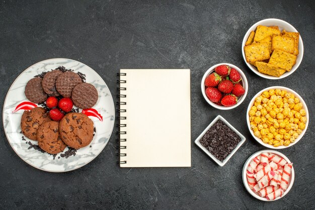 Vista superior del delicioso plato de bizcocho con trozos de fresas frescas de chocolate y tazones de galletas cariño sobre fondo negro con espacio libre