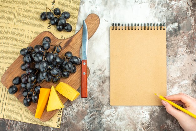 Vista superior del delicioso racimo de uva negra fresca y varios tipos de queso en la tabla de cortar de madera y un cuchillo junto al cuaderno sobre fondo de colores mezclados