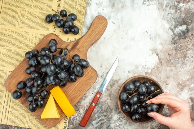 Vista superior del delicioso racimo de uva negra fresca y varios tipos de queso en la tabla de cortar de madera y en un cuchillo marrón sobre fondo de colores mezclados