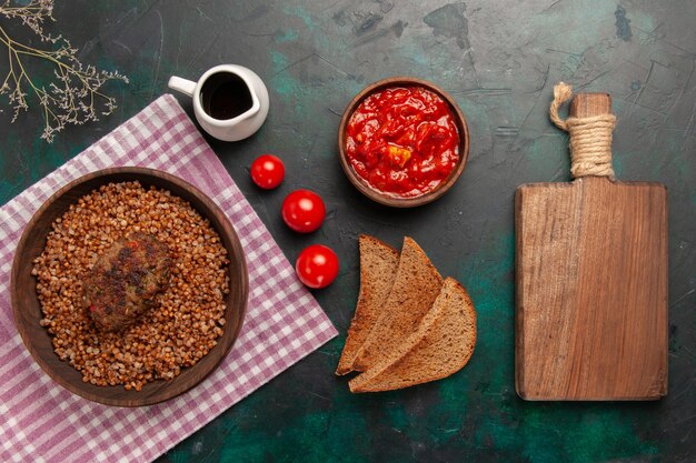 Vista superior delicioso trigo sarraceno cocido con hogazas de pan oscuro y chuleta en el plato de verduras de comida de ingrediente de superficie verde oscuro