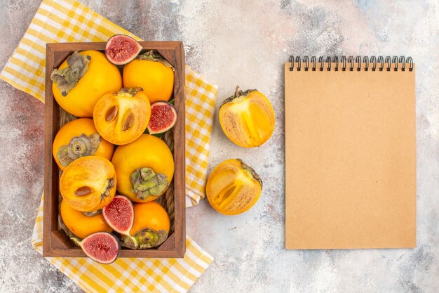 Vista superior deliciosos caquis e higos cortados en caja de madera toalla de cocina amarilla un cuaderno sobre fondo desnudo