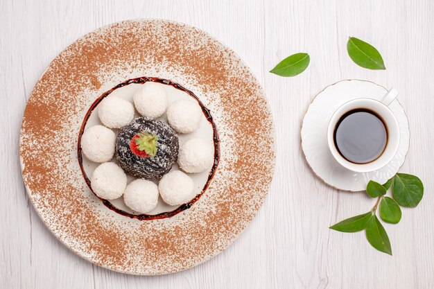 Vista superior de deliciosos caramelos de coco con pastel de chocolate y una taza de té sobre fondo blanco, pastel de té, galletas, dulces, galletas dulces