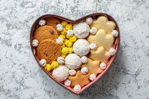 Vista superior de deliciosos dulces, galletas, galletas y caramelos dentro de una caja en forma de corazón sobre una superficie blanca, pastel de azúcar, té, dulce y delicioso