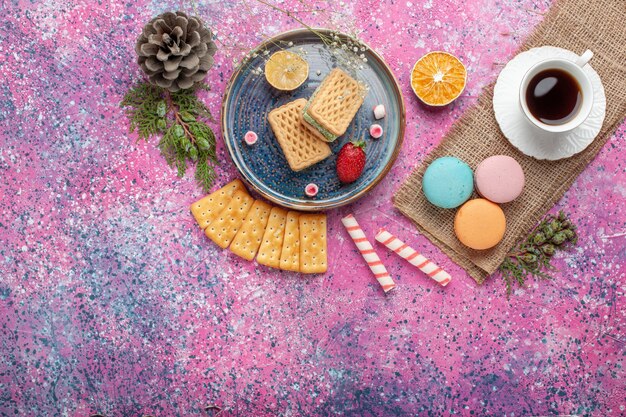 Vista superior de deliciosos gofres con macarons franceses y té en la superficie rosa