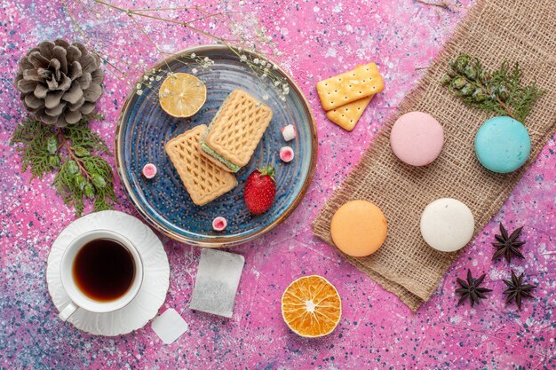 Vista superior de deliciosos gofres con macarons y taza de té en la superficie rosa