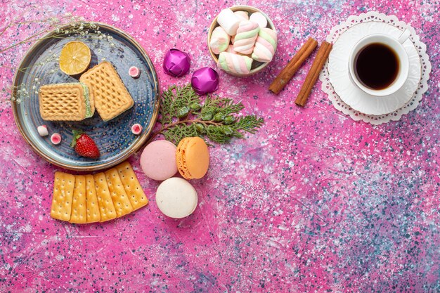 Vista superior de deliciosos gofres con macarons y taza de té en la superficie rosa