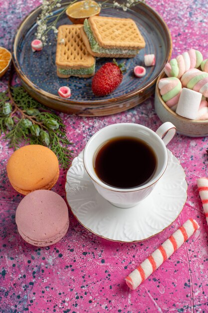 Vista superior de deliciosos gofres con taza de té macarons y malvaviscos en superficie rosa