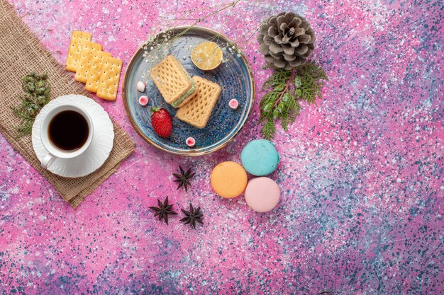 Vista superior de deliciosos gofres con taza de té y macarons en superficie rosa