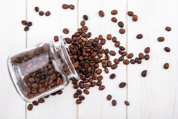 Vista superior de deliciosos granos de café cayendo de un frasco de vidrio sobre un fondo blanco de madera