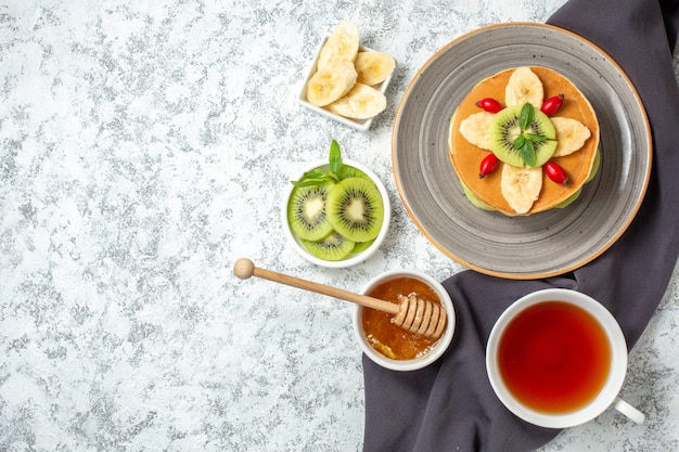 Vista superior deliciosos panqueques con frutas en rodajas y una taza de té en la superficie blanca, postre dulce, azúcar, desayuno, tortas de color