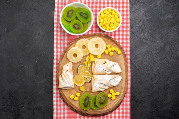 Vista superior de deliciosos pasteles con anillos de piña secos y kiwis en un espacio gris oscuro