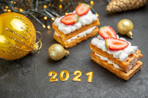 Vista superior de deliciosos pasteles de crema con fresas alrededor de juguetes de árbol de año nuevo sobre fondo gris