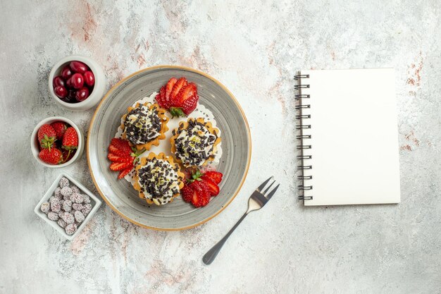 Vista superior de deliciosos pasteles con crema y fresas en la celebración de cumpleaños de escritorio blanco pastel de galletas dulces