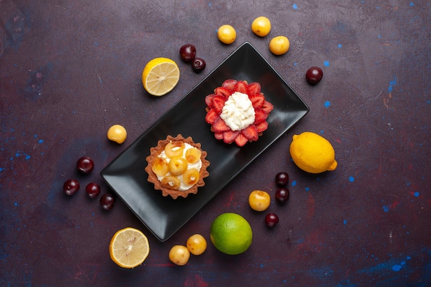 Vista superior de deliciosos pasteles cremosos dentro de la placa con limones y frutas sobre una superficie oscura