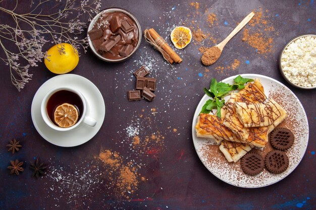 Vista superior de deliciosos pasteles dulces con galletas de chocolate y una taza de té en el espacio oscuro