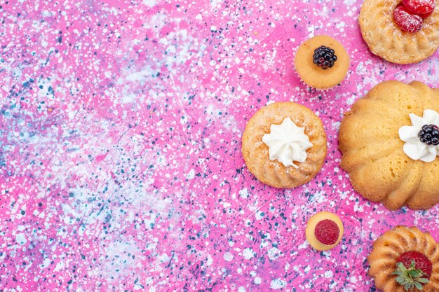Vista superior de deliciosos pasteles horneados con crema junto con bayas en el escritorio de color púrpura brillante, pastel de baya de galleta dulce hornear té