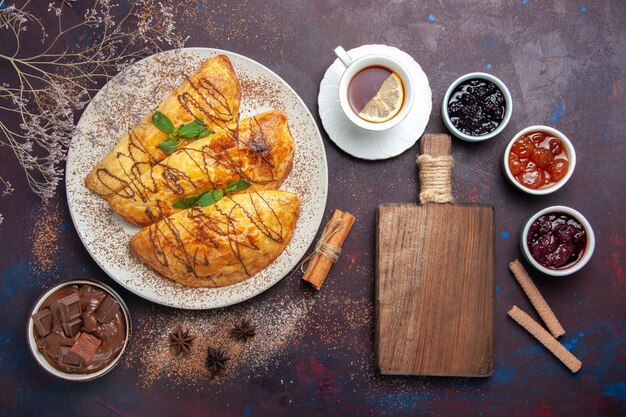 Vista superior de deliciosos pasteles horneados con taza de té y mermelada en el escritorio oscuro