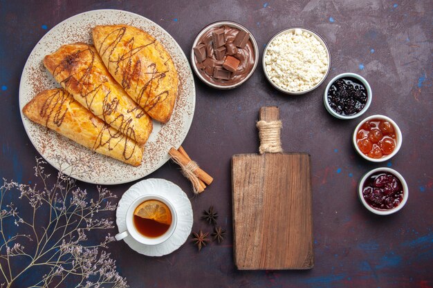 Vista superior de deliciosos pasteles horneados con taza de té y mermelada en el escritorio oscuro