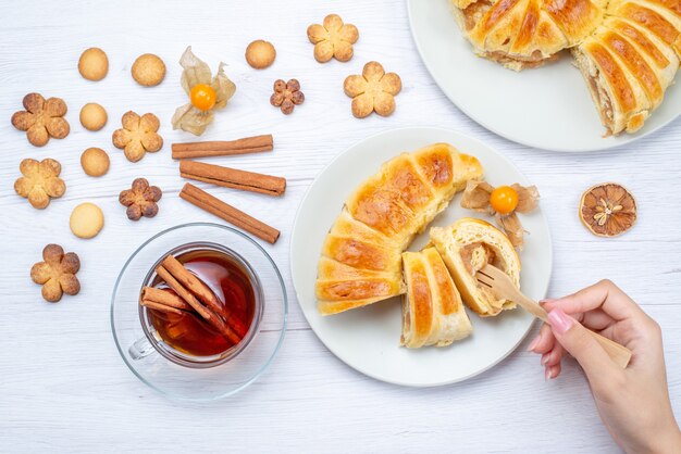 Vista superior deliciosos pasteles en rodajas dentro de la placa con relleno junto con té, canela y galletas en el escritorio ligero, pastelería, galleta, galleta, dulce