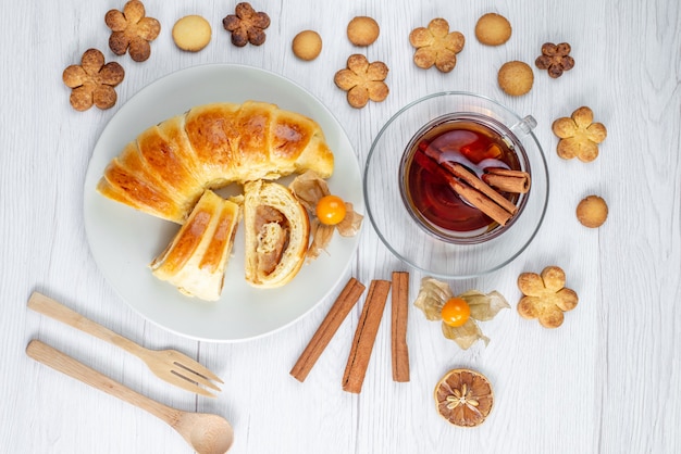 Vista superior de deliciosos pasteles en rodajas dentro de la placa con relleno junto con té y galletas en el escritorio blanco, pastelería galleta galleta dulce