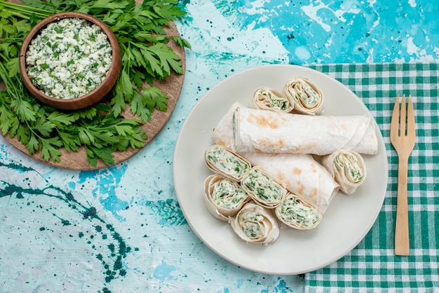 Vista superior de deliciosos rollos de vegetales enteros y en rodajas con verduras y ensalada en el escritorio azul, bocadillo de vegetales de rollo de comida