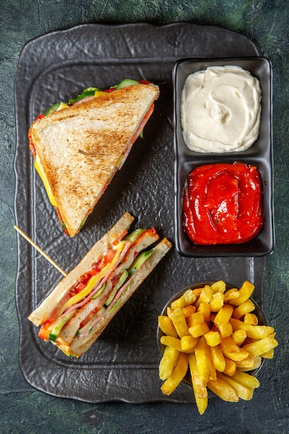 Vista superior deliciosos sándwiches de jamón con papas fritas y condimentos superficie oscura
