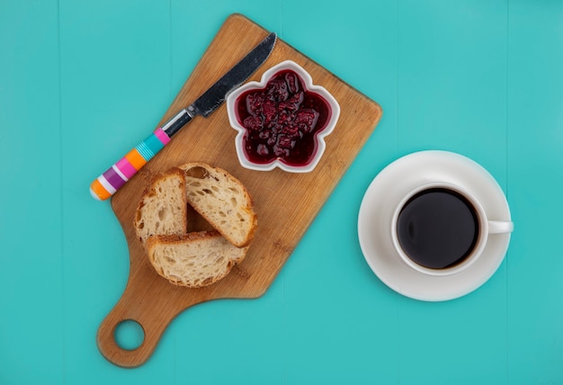 Vista superior del desayuno con baguette en rodajas y mermelada de frambuesa con un cuchillo en la tabla de cortar y una taza de té sobre fondo azul.