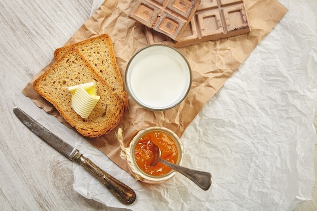 Vista superior del desayuno con chocolate, mermelada, pan tostado seco, mantequilla y leche. Todo en papel artesanal y cuchillo y cuchara vintage con pátina.