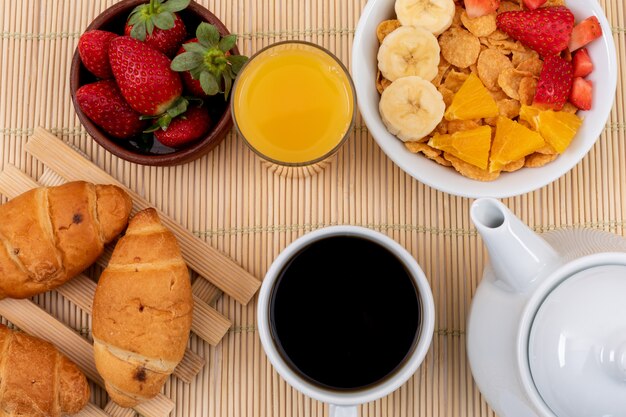 Vista superior del desayuno con copos de maíz, fresas, jugo y cruasanes en superficie horizontal de paja