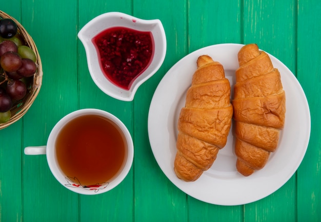 Vista superior del desayuno con croissants en un plato taza de té mermelada de frambuesa en un tazón y una canasta de endrinas de uva sobre fondo verde